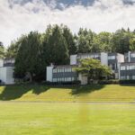 Row of modern, townhouse-style student housing buildings surrounded by lush green trees and a well-maintained grassy field, located on the BCIT Burnaby campus under a partly cloudy sky.